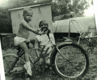 I spot Boo Boo as he “rides” my broken-down bike. Boatbuilding backdrop.