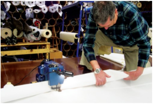 The sail must be rolled so it will pass inside the “throat” of the sewing machine. Putting a portable machine on the floor provides, in essence, a large working table. The foot pedal for the machine is under Jim’s right foot.