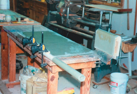 On Paul’s workbench are the three components from which the waterlift muffler was built: on top of a waxed plate-glass mirror, he laid up a sheet of fiberglass for the sides, top, bottom, and ends. The fiberglass tube, made by winding epoxy-saturated fiberglass cloth around a PVC pipe, became the inlet, outlet, and crossover tubes. He also laid up — over a wooden male mold — the valley to allow the propeller shaft to pass through the waterlift. That assembly is to the right.