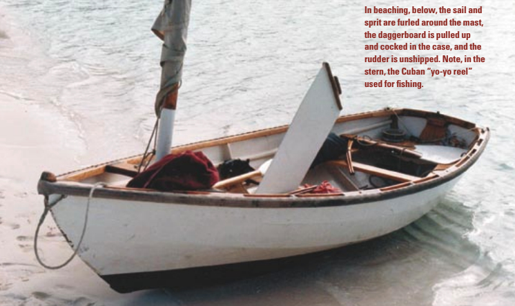 In beaching, below, the sail and sprit are furled around the mast, the daggerboard is pulled up and cocked in the case, and the rudder is unshipped. Note, in the stern, the Cuban “yo-yo reel” used for fishing.