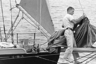 Al Petersen stands on footropes with his knees securely around the bowsprit. Note how Al has tied off the staysail boom to keep it out of mischief while he is nearby. This is the bow of Stornoway, the 33-foot cutter that Al sailed around the world.