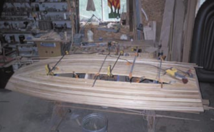 The stripping coming to a conclusion on the starboard side, below. John notes, “I like to do as many straight strips as I can. When they start to curve too much, I start at the opposite side and work up or down toward the others. When they meet, I alternate up and down as the space gets smaller. The strips are shorter, and you can use the cutoffs and short pieces to fill in. Just make sure that you do the same pattern on both sides of the boat for a balanced look. This is like laying tile. You can do it messy or create a mosaic pattern with different wood that illustrates the history of the world, if you want. The wood strips are there to hold up the fiberglass cloth.” When the stripping is done, he points out, “It’s time for filling and sanding and sanding and sanding.” The builder does not need to get overly fussy with filling gaps and holes. This does not affect the structural and waterproof nature of the hull. “However,” John says, “I prefer not to see too much light coming through my hull.”